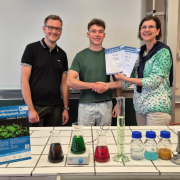 Fachraum Chemie, hinter dem gefliesten Pult stehen ein Chemielehrer, ein Schüler aus der 12 und eine Schulleiterin. Alle drei schauen fröhlich drein.