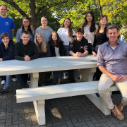 An einer Outdoor-Sitzgruppe aus Beton sitzen und stehen 10 Schülerinnen und zwei Lehrer.