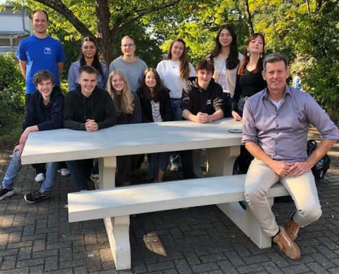 An einer Outdoor-Sitzgruppe aus Beton sitzen und stehen 10 Schülerinnen und zwei Lehrer.