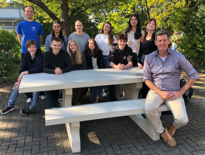 An einer Outdoor-Sitzgruppe aus Beton sitzen und stehen 10 Schülerinnen und zwei Lehrer.