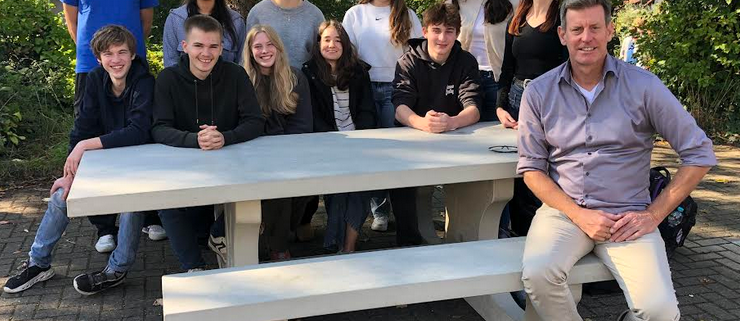 An einer Outdoor-Sitzgruppe aus Beton sitzen und stehen 10 Schülerinnen und zwei Lehrer.