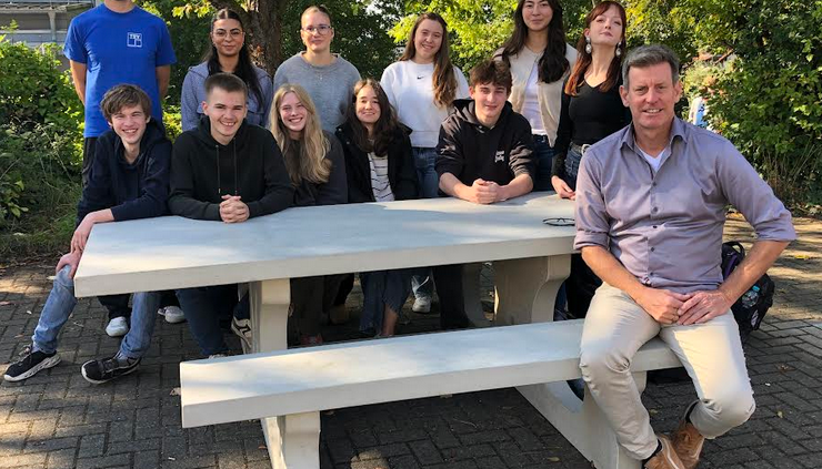 An einer Outdoor-Sitzgruppe aus Beton sitzen und stehen 10 Schülerinnen und zwei Lehrer.