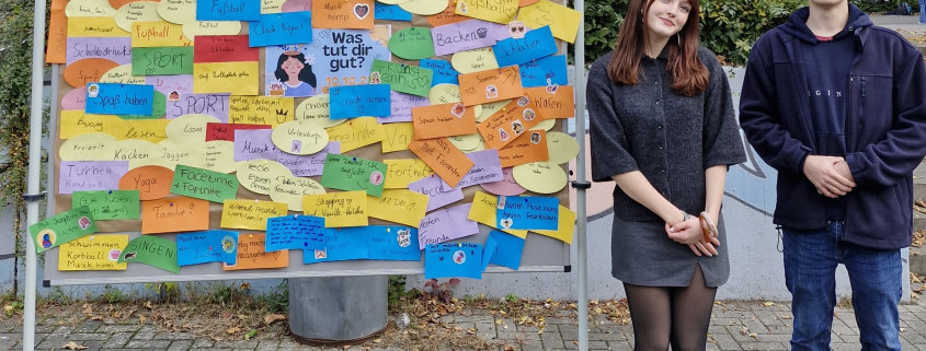 Das Schülersprecherteam steht neben einer Stellwand auf dem Schulhof.
