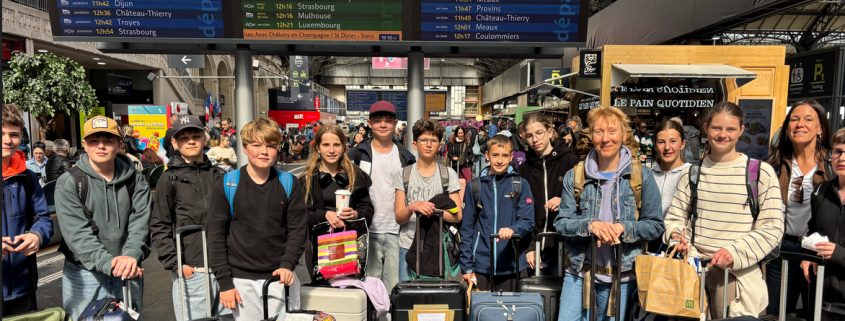 eine Gruppe von 15 Schülerinnen und Schülern mit Gepäck wartet in einem französischem Bahnhof